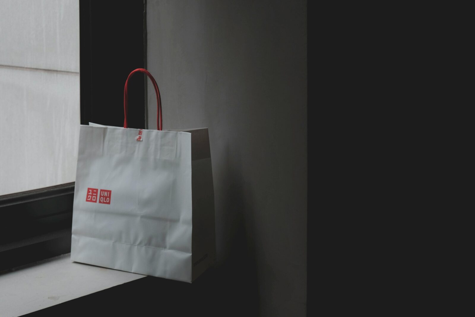 A white paper shopping bag with red handles and a logo, placed on a windowsill in a minimalistic setting.
