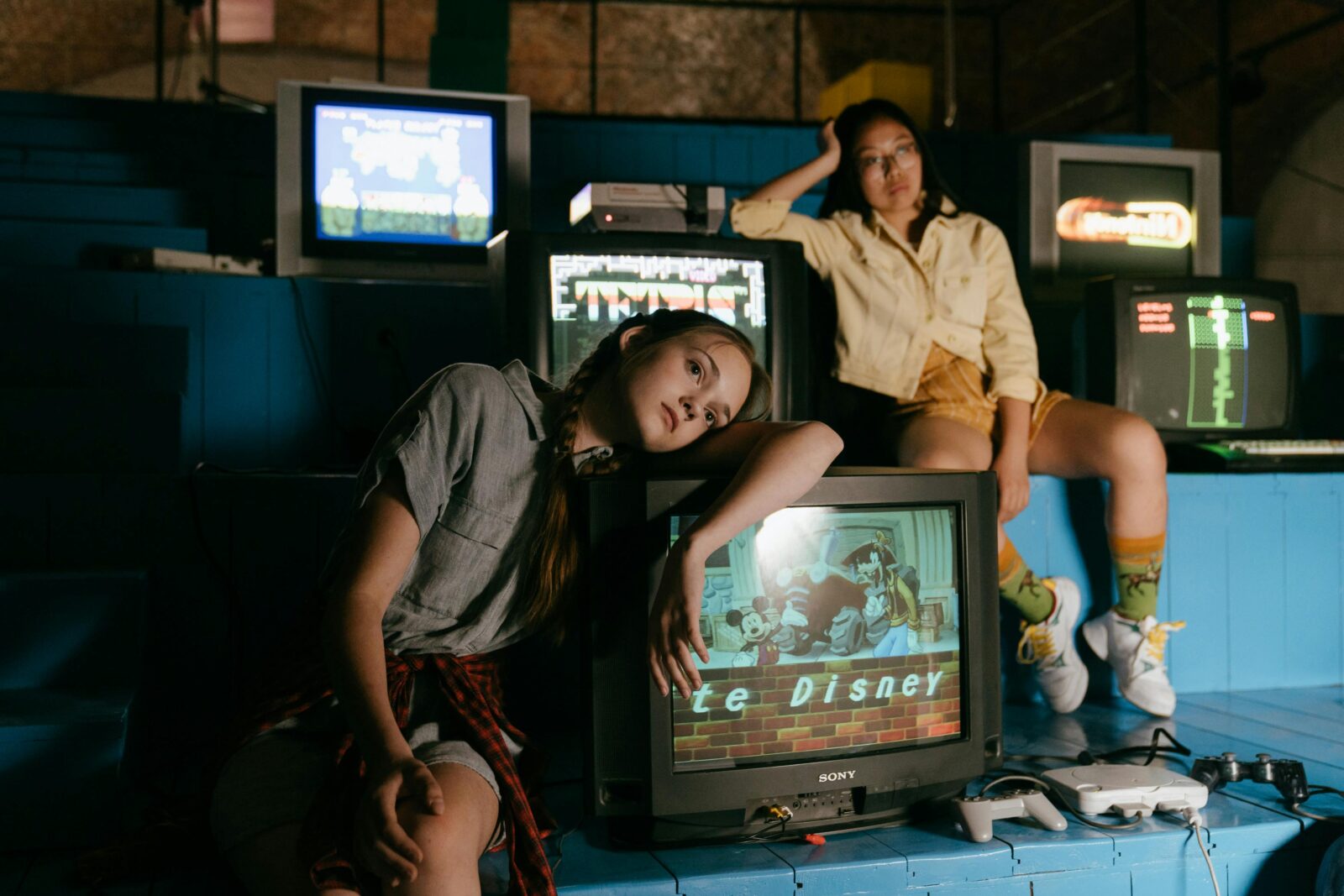 Two women in casual outfits immersed in retro gaming with vintage televisions and game consoles.